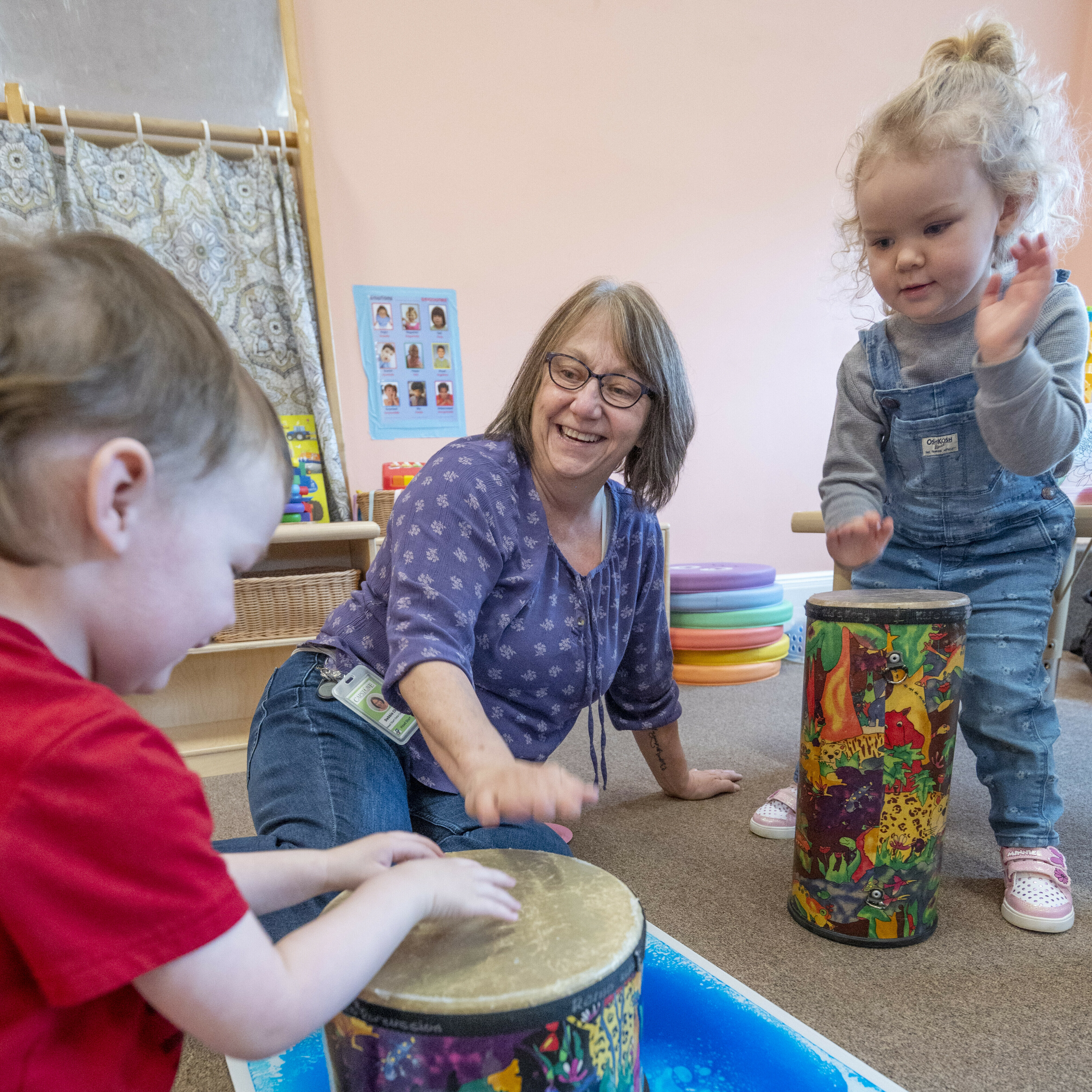 Children and a caregiver play drums and smile in a colorful classroom setting.
