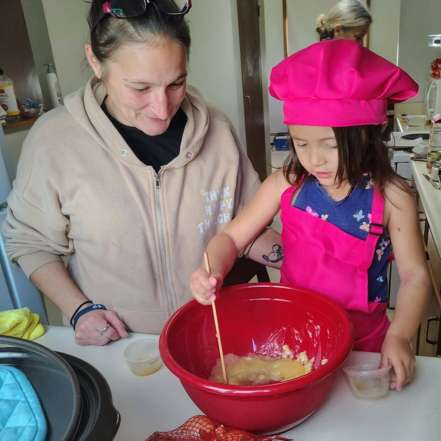 A woman watches as a girl in a pink chef hat stirs a mixture in a red bowl.