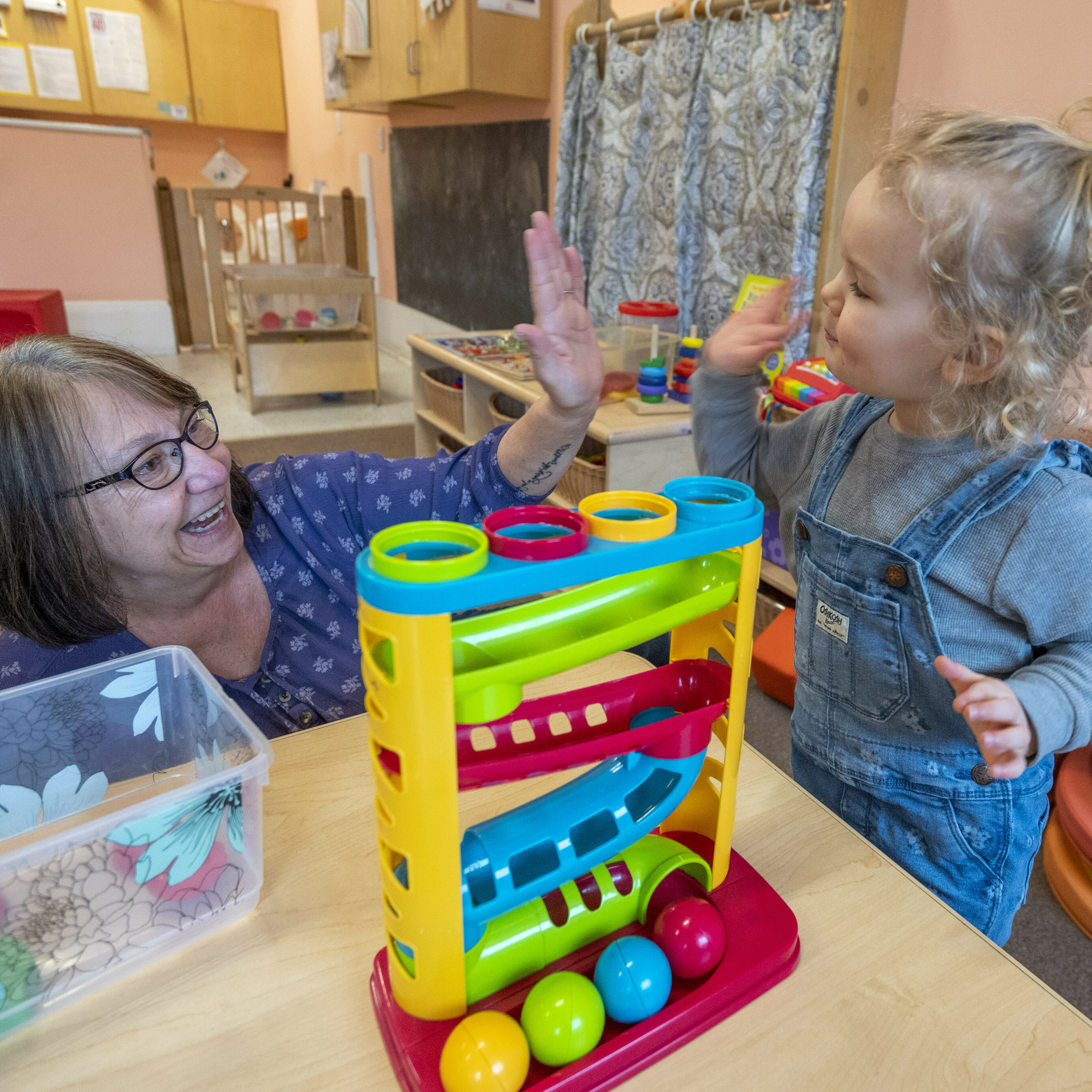 Child and woman happily interact, playing with colorful toy in a bright classroom.