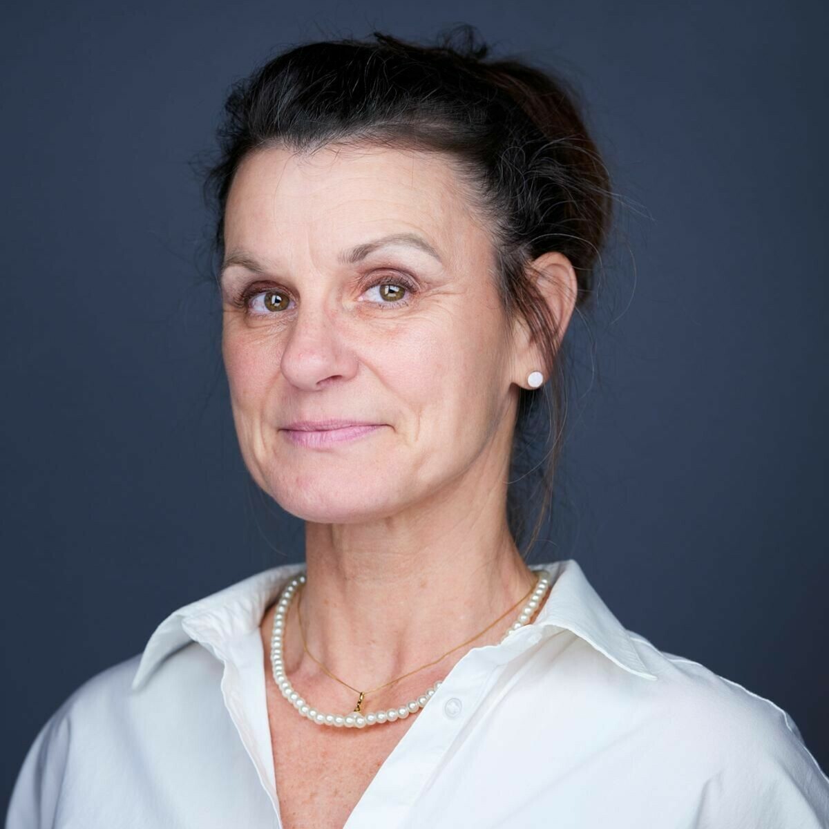 A smiling woman with brown hair, wearing a white shirt and pearl necklace, against a dark background.