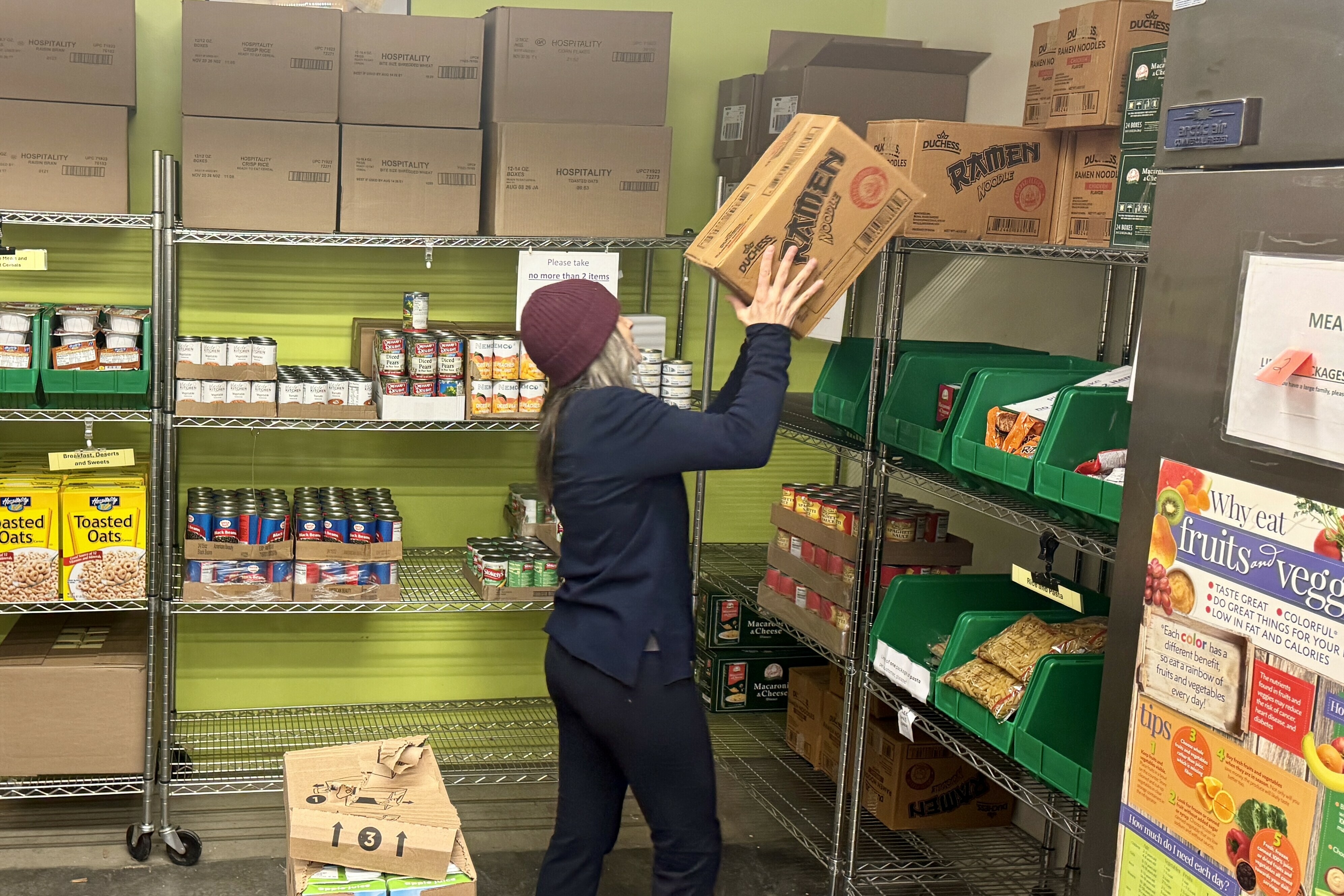 A person shelves boxes of ramen noodles in a food pantry with various canned goods.