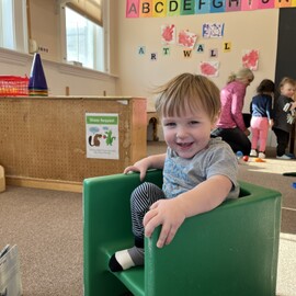 A child sitting in a green chair smiles, while other kids play in the background.