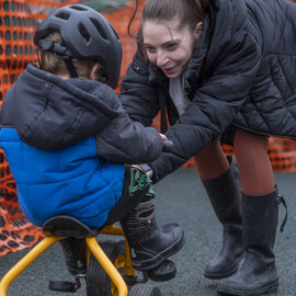 A child in a helmet rides a tricycle while an adult assists them, smiling.