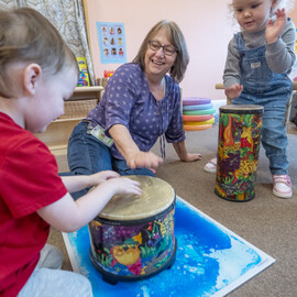 Children play drums with a smiling adult in a colorful classroom setting.