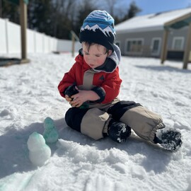 Child in winter gear sitting in snow, creating snow figures with colored spray.