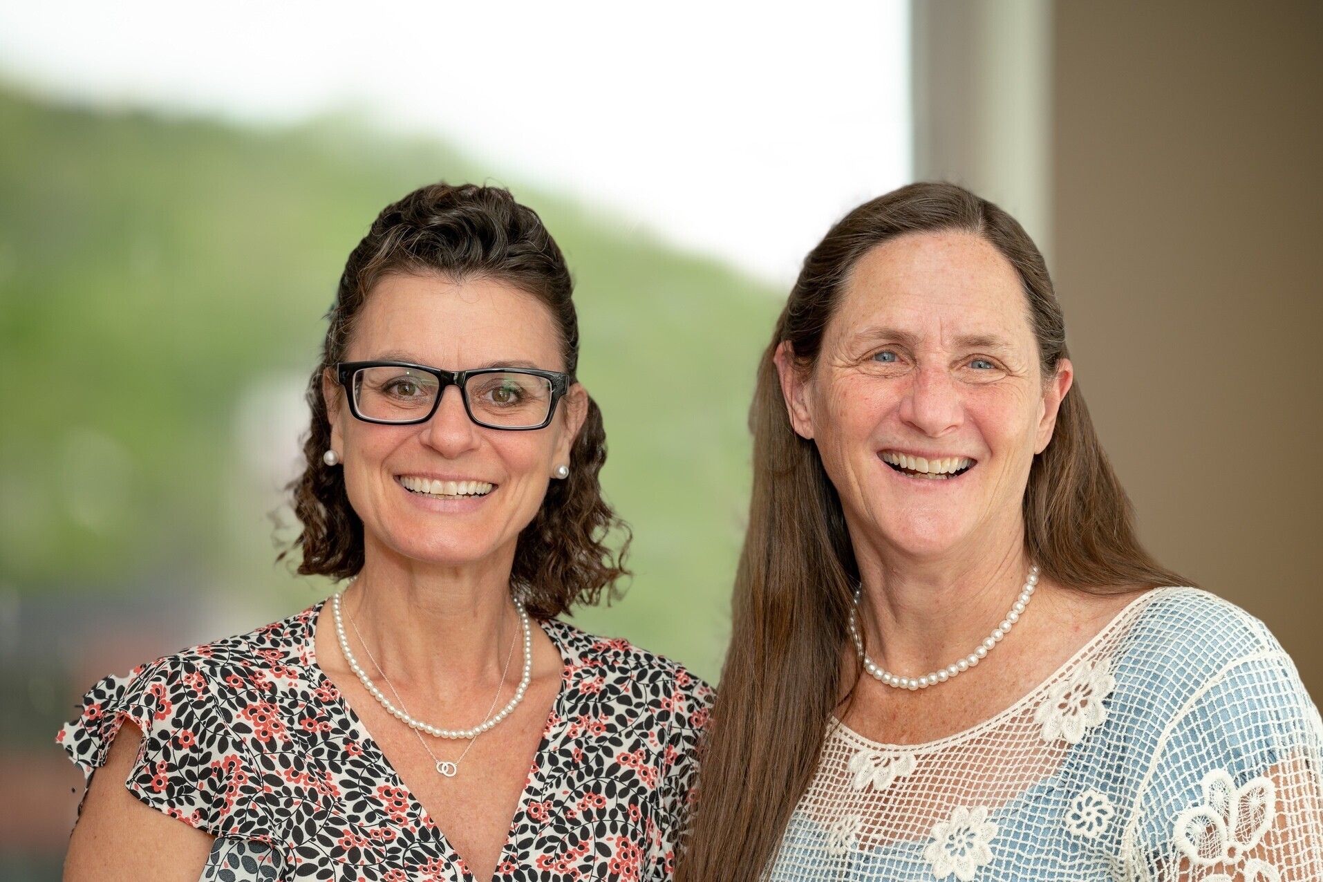 Two smiling women look towards the camera. Rosie, on the left has shoulder length dark brown hair and dark framed glasses. Mary, on the right, has long light brown hair and is wearing a pale blue shirt with crocheted overlay.