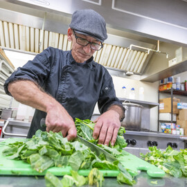 Chef chopping green leafy vegetables on a cutting board in a kitchen.